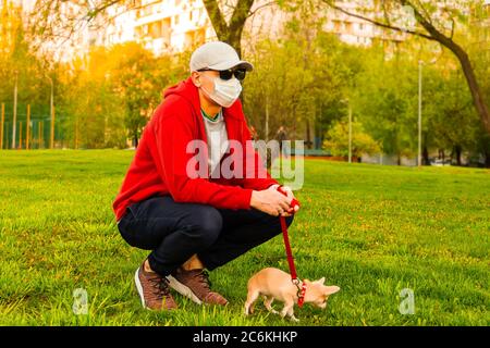 Ritratto di giovane caucasico in felpa rossa, maschera medica di protezione e occhiali da sole rimanere nel parco a giorno di sole. Papà camminare il simpatico cane cucciolo ch Foto Stock