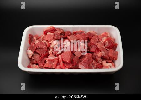 Carne grezza tagliata a cubetti in un contenitore bianco di polistirolo isolato su fondo nero con spazio per la copia del testo Foto Stock