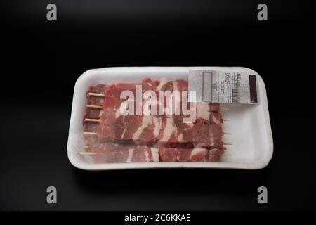 Carne grezza tagliata a cubetti su spiedini di legno in un contenitore bianco di polistirolo con codice a barre, isolato su fondo nero con spazio di copia fo Foto Stock