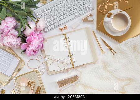 Diario e cancelleria dorata per donne. Bouquet di peonie rosa. Bicchieri, una tastiera bianca, una penna, forbici e caffè sulla scrivania. Foto Stock