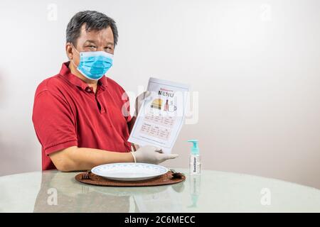 Concettuale dell'uomo asiatico con maschera facciale, guanto e disinfettante che guarda al menu usa e getta al ristorante, come parte del nuovo stile di vita normale. Foto Stock