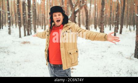 Bell'uomo in piedi e che canta in foresta nella stagione invernale. Giovane maschio che indossa una giacca beige sopra una felpa con cappuccio rossa e un cappello con le braccia Foto Stock