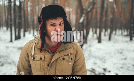 Uomo grimaces in foresta nella stagione invernale. Ritratto di pazzo maschio che indossa copricapo e grimacing faccia da vicino nella foresta invernale. Foto Stock