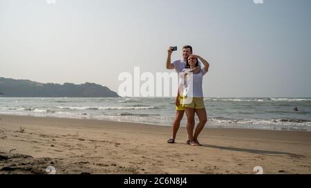 Felice coppia che prende selfie vicino al mare. Coppia amorosa che si abbraccia durante la data sulla spiaggia contro il mare ondulato e cielo senza nuvole. Foto Stock