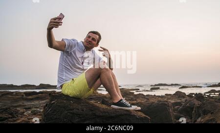 Uomo rilassato seduto sulla pietra e prendendo selfie su smartphone a riva. Vista laterale di un uomo piacevole che trascorre del tempo godendo la vacanza e fotografando Foto Stock