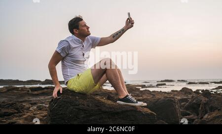 Uomo rilassato seduto sulla pietra e prendendo selfie su smartphone a riva. Vista laterale di un uomo piacevole che trascorre del tempo godendo la vacanza e fotografando Foto Stock