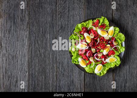 insalata di polpo con uova sode, capperi, cipolla marinata su foglie di lattuga su un piatto nero su un tavolo di legno scuro, vista orizzontale da abov Foto Stock