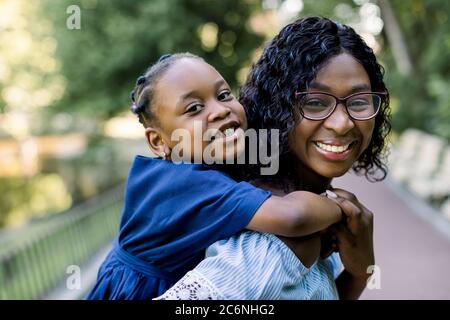 Felice madre africana con pelle scura che tiene il suo bambino in un parco. Sorridente giovane madre allegra che porta la sua piccola figlia carina sul piggyback e avendo Foto Stock