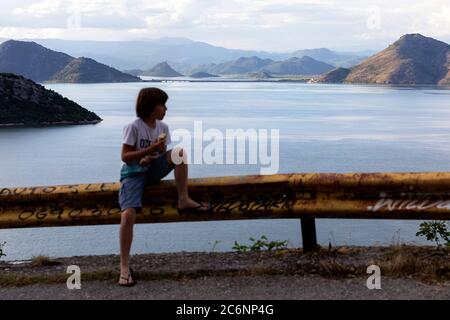 Ragazzo seduto sulla recinzione della strada con uno spuntino con una bella vista sul Lago Skadar e Virpazar ponte al tramonto, Montenegro Foto Stock
