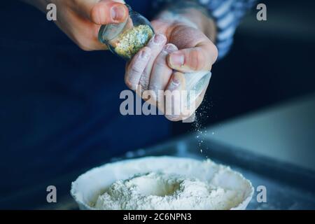 Una cuoca in un grembiule blu tiene un bicchiere di sale shaker nelle mani macchiate di farina e versa il condimento nella farina in un recipiente per l'impasto. Il processo di co Foto Stock