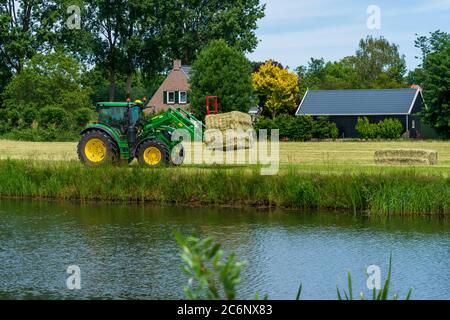 Dordrecht, Paesi Bassi - 1 giugno 2020: Trattore verde che carica balle di fieno su un rimorchio nella campagna rurale. Il Parco Nazionale di Biesbosch è uno dei più grandi parchi nazionali dei Paesi Bassi Foto Stock