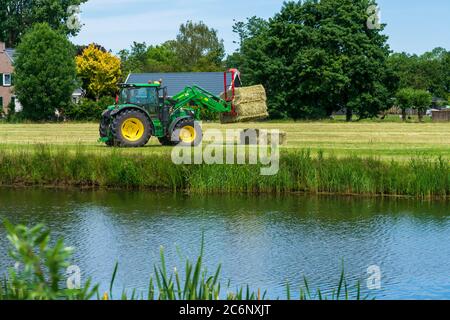 Dordrecht, Paesi Bassi - 1 giugno 2020: Trattore verde che carica balle di fieno su un rimorchio nella campagna rurale. Il Parco Nazionale di Biesbosch è uno dei più grandi parchi nazionali dei Paesi Bassi Foto Stock