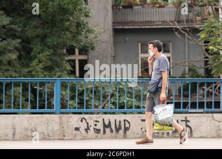 Belgrado, Serbia - 3 luglio 2020: Un uomo che indossa una maschera chirurgica a piedi lungo la strada con borsa della spesa accanto all'edificio residenziale in estate Foto Stock