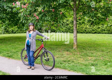 donna incinta sul prato verde in un parco che tiene una bicicletta pronta a correre. Foto Stock