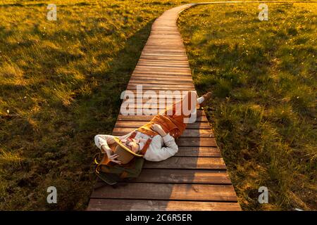 Donna naturalista in cappello beige e tute arancioni poggianti su uno zaino su un percorso di legno attraverso palude torba nel parco nazionale della fauna selvatica a sole Foto Stock