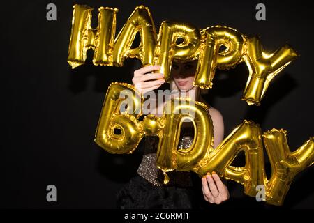 Giovane donna elegante che tiene palloncini a forma di lettera di colore dorato mentre si congratulano per il vostro compleanno contro sfondo nero Foto Stock