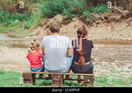 una famiglia con bambini si siede sulla riva del fiume su una panchina che abbrancava Foto Stock