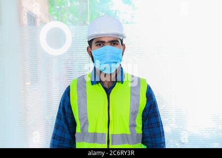 Giovane ingegnere che indossa maschera, closeup di barba uomo che indossa camicia blu con giubbotto giallo e casco ywhite, torna al lavoro dopo le estremità di blocco a causa di covid Foto Stock