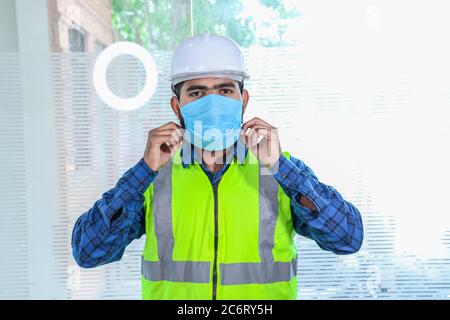 Giovane ingegnere che indossa una maschera di regolazione sorridente, primo piano di barba uomo che indossa una camicia blu con gilet giallo e casco bianco, torna al lavoro dopo il blocco Foto Stock