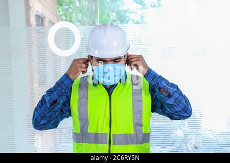 Giovane ingegnere che indossa una maschera di regolazione sorridente, primo piano di barba uomo che indossa una camicia blu con gilet giallo e casco bianco, torna al lavoro dopo il blocco Foto Stock