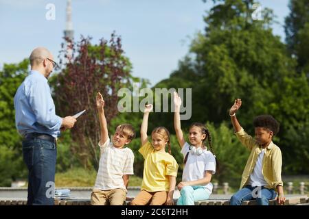 Gruppo multietnico di bambini allegri che alzano le mani e ascoltano l'insegnante di sesso maschile mentre si gode una lezione all'aperto alla luce del sole, spazio di copia Foto Stock