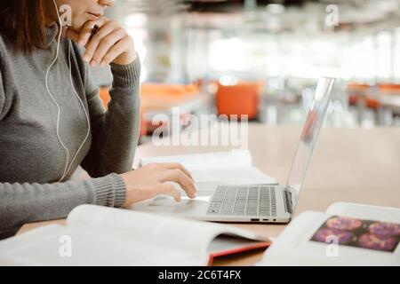 Una ragazza studentessa con le cuffie che si prepara all'esame e utilizza il computer portatile mentre si siede al tavolo presso la biblioteca universitaria. Foto Stock