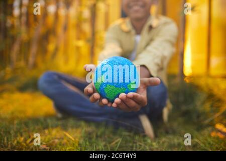 Primo piano di un ragazzo afroamericano irriconoscibile che tiene il pianeta in mano mentre si siede su erba verde all'aperto alla luce del sole, copia spazio Foto Stock