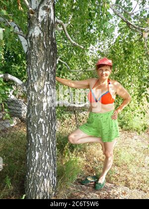 Primo piano ritratto di una donna anziana sorridente che è divertente posare vicino recinzione in cortile sotto l'albero di betulla. Foto Stock