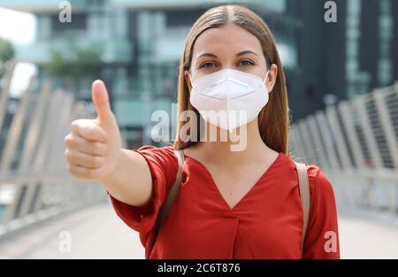 COVID-19 donna di affari ottimista che indossa la maschera protettiva KN95 FFP2 mostrando pollici in su nella strada moderna della città Foto Stock