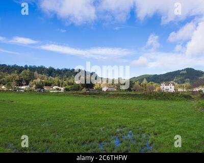 Fattoria e vecchia villa nel villaggio di Sete Cidades con erba verde lussureggiante e acqua puddle. Colline con boschi e pascoli, cielo blu e nuvole bianche Foto Stock