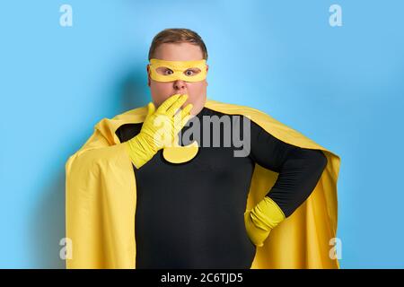 il giovane caucasico scioccato fat superman guarda la macchina fotografica, sta chiudendo la bocca con le mani in guanti protettivi. mr soggiorno giusto sorpreso dopo aver visto dir Foto Stock