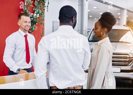 amichevole uomo caucasico amministratore e consulente di concessionaria accoglie i clienti, la gente nera è venuto a fare l'acquisto, per acquistare la migliore auto Foto Stock