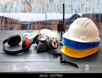 Attrezzatura di protezione per la sicurezza sul lavoro. Indumenti protettivi industriali su tavolo in legno, sfocatura sfondo cantiere. Hardhats, cuffie, maschera respiratoria, Foto Stock