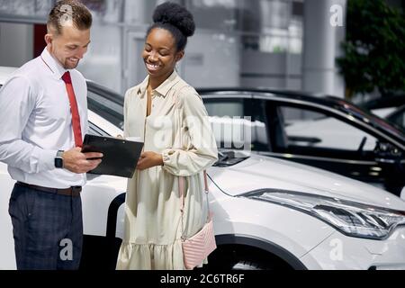 ritratto di venditore e cliente africano donna che hanno conversazione in showroom auto. bel venditore caucasico offre buone condizioni di vendita Foto Stock