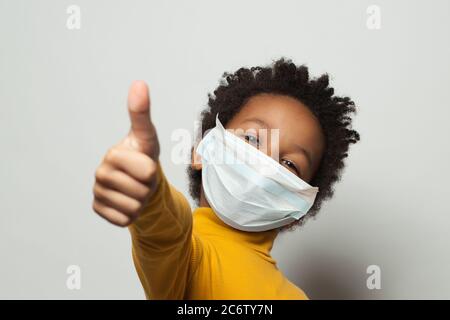 Felice Africano americano nero capretto in maschera medica protettiva del viso mostrando pollice su bianco Foto Stock