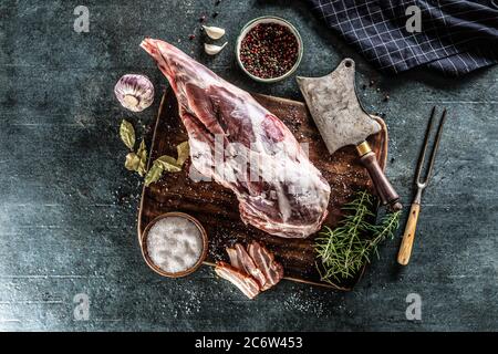 Coscia di agnello cruda circondata da sale, pepe, aglio, pancetta e rosmarino su una superficie di vinateg Foto Stock