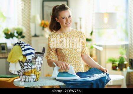 moderna casalinga di 40 anni in abito giallo con cesto di vestiti lavati e ferro e asse da stiro nel moderno soggiorno in giornata di sole. Foto Stock