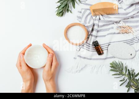 Mani femmina piatto tenere il vaso crema idratante accanto al tovagliolo e zero rifiuti accessori bagno con rami verdi su tavolo bianco, vista dall'alto. Foto Stock