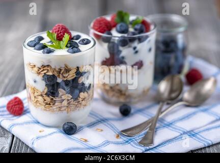 Granola con yogurt bianco con lamponi e mirtilli in due bicchieri su tovagliolo blu. Foto Stock
