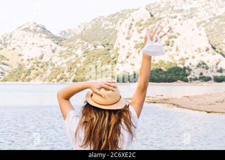 Una donna, adolescente in un cappello che tiene una maschera di fronte ad un lago nella nuova normalità. Concetto di libertà e di uscire in natura. Foto Stock