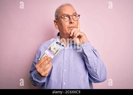 Uomo di mezza età che tiene un mazzo di banconote di dollari su sfondo rosa isolato faccia seria pensare alla domanda, idea molto confusa Foto Stock
