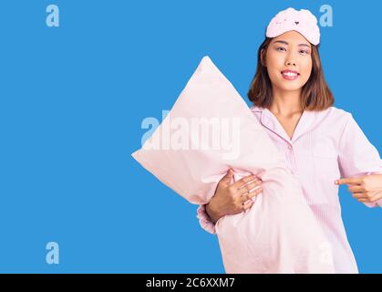 Giovane bella ragazza cinese che indossa la maschera di sonno e pajama tenendo cuscino sorridente felice di puntare con mano e dito Foto Stock