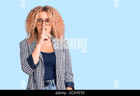 Giovane bionda con capelli ricci che indossa giacca da lavoro e occhiali che chiedono di essere tranquilla con le dita sulle labbra. Silenzio e concetto segreto. Foto Stock