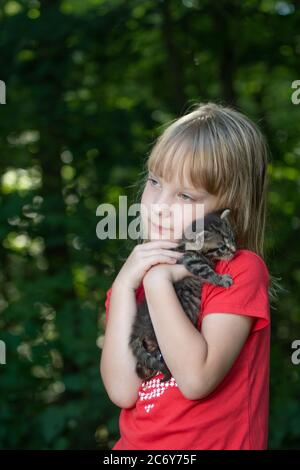 Una ragazza di cinque anni che tiene un gattino tabby all'aperto in estate con alberi sullo sfondo. Foto Stock