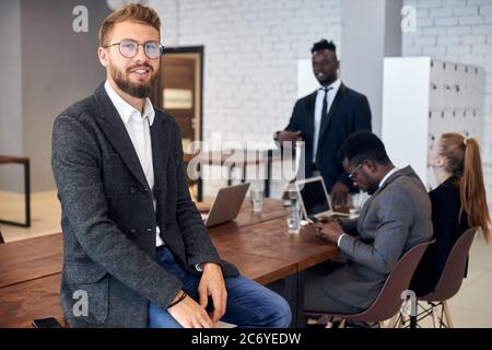 Uomo attraente che guarda con sicurezza la fotocamera mentre i suoi partner commerciali hanno una discussione in ufficio Foto Stock