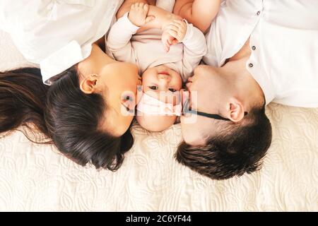 Felice giovane padre, madre e simpatico bambino ragazzo sdraiato su letto leggero. Madre e padre baciano bambino. Foto Stock