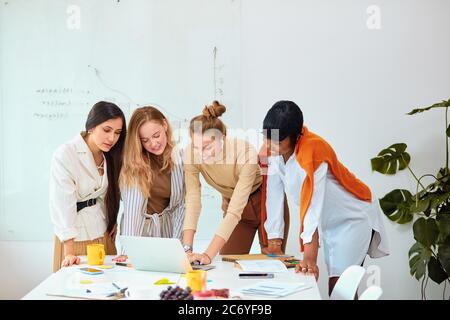 Donne caucasiche e africane, le donne di affari si sono riunite in un moderno ufficio bianco per discutere le idee di affari usando il laptop, lavoro di squadra di belle Signore indossare Foto Stock