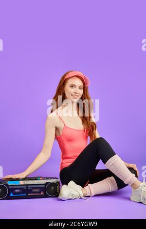 bella donna fitness con pelle incandescente, fracelle e capelli ricci rossi, si siede sul pavimento con la mano sul lettore audio portatile, sorride gioiosamente, soddisfacente Foto Stock