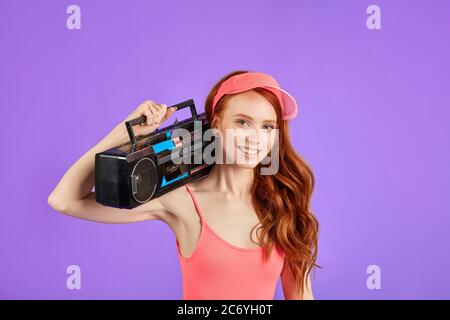 giovane donna dai capelli rossi con frettoloni guarda in macchina fotografica con sorriso, tiene stereo e cassetta lettore di nastri sulla spalla, indossa un cappello rosa e aerobica Foto Stock