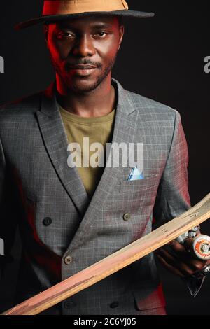Giovane bel uomo d'affari africano che indossa abito grigio e cappello elegante in posa con longboard isolato contro sfondo rosso scuro fumoso Foto Stock